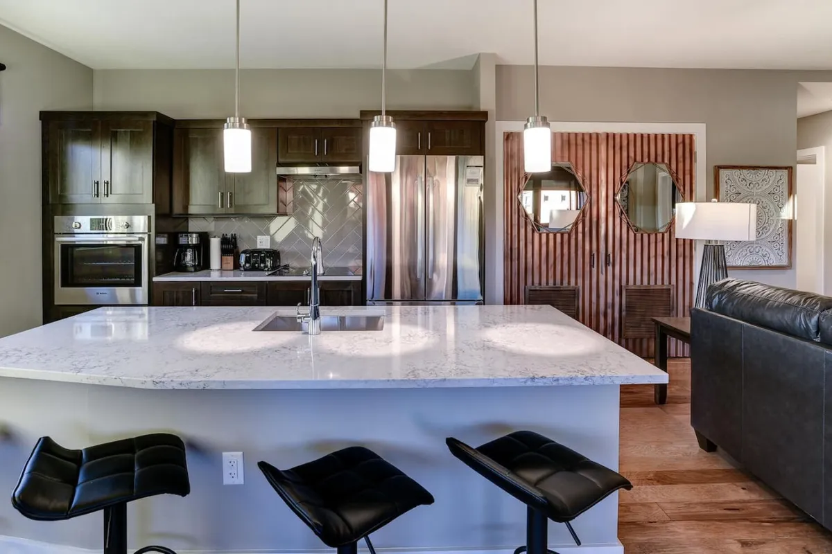 A bright, modern kitchen with a kitchen island, pendant lights, dark cabinetry, stainless steel appliances, and mirrored decor. The layout suggests a space for preparing and sharing meals with friends or family, and the design is both stylish and welcoming.