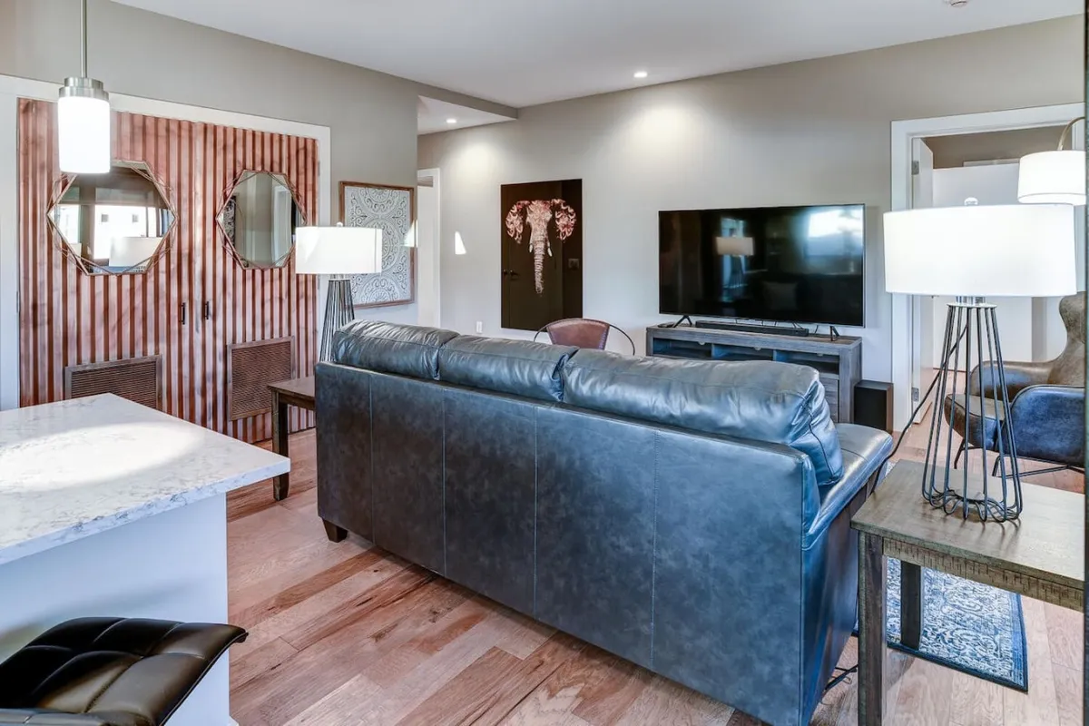 A cozy, modern living area with a leather sofa, TV unit, wooden accent wall, decorative mirrors, and table lamps. The space is inviting and well-lit, perfect for unwinding after a day of exploring Asheville or sharing stories with friends in a communal setting.
