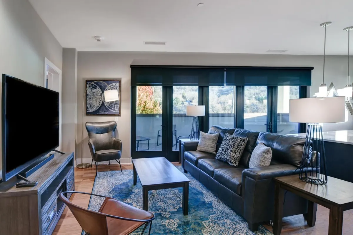 A cozy living room featuring a plush sofa, coffee table, modern art on the walls, and a television. Large windows let in abundant natural light, creating a bright and inviting space. The arrangement encourages relaxing, socializing, and taking in the urban energy outside.