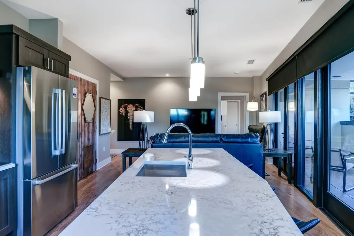 A contemporary kitchen layout featuring a marble-topped island, stainless steel refrigerator, and a comfortable blue leather sofa in the background, with a TV mounted on the wall nearby. The arrangement hints at easy transitions between cooking, dining, and relaxed socializing.