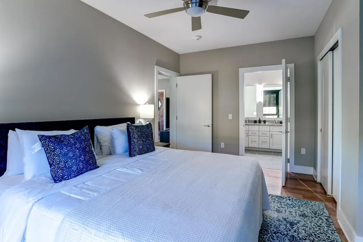 This image displays a tranquil bedroom with decorative pillows, a large mirror in the bathroom visible nearby, and a stylish light fixture next to the bed. The interplay of light and space suggests an inviting haven to return to after a day of exploration.