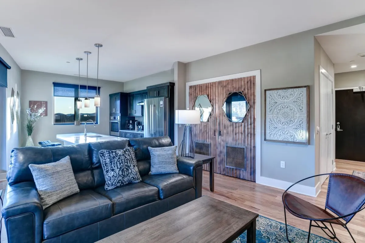 An open-plan living and dining area with a comfortable sofa adorned with decorative pillows, a wooden front door, a visible kitchen area, and stylish wall art. A dining table is set up for gatherings, blending relaxation and sociability.