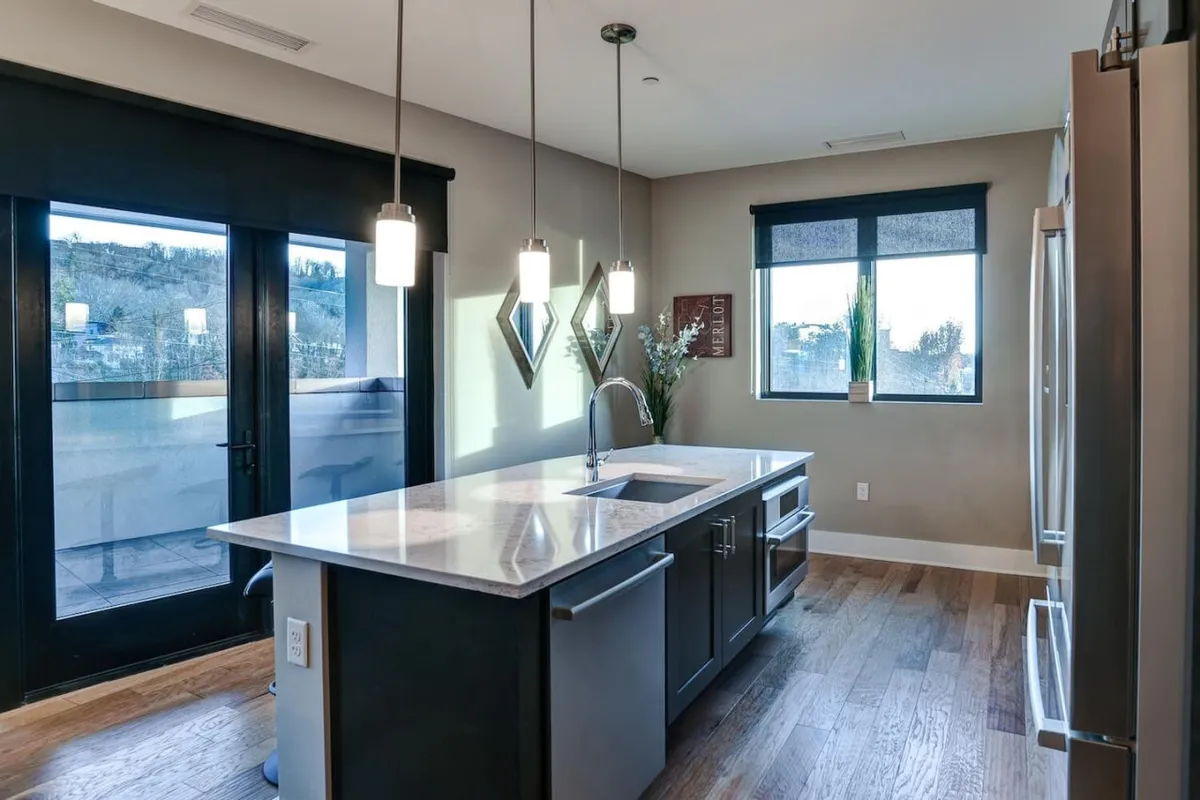 A bright, modern kitchen featuring a spacious island with a sink, sleek pendant lighting overhead, and a large window that provides a view of the city lights. The space is open and airy, inviting both culinary creativity and social interaction.