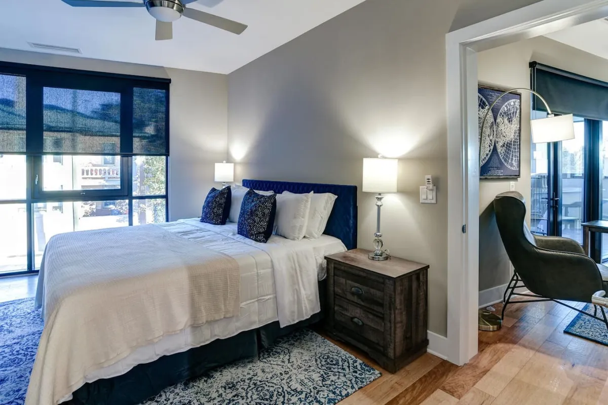 This image features a cozy bed adorned with decorative pillows, a nightstand with a softly glowing lamp, and a window that floods the room with natural light. The view outside the window and the inviting ambiance evoke a sense of peaceful mornings and relaxation.
