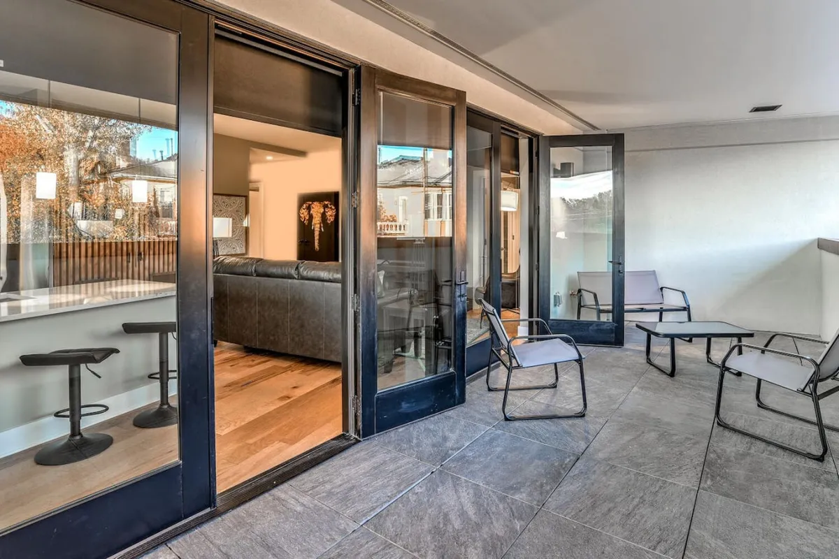 The image showcases the condo’s spacious enclosed balcony with comfortable outdoor seating, accessible through sliding glass doors. The sun sets over downtown Asheville and the surrounding mountains, visible from the balcony. Through the glass, you can see a glimpse of the inviting living room, creating a seamless indoor-outdoor living experience.