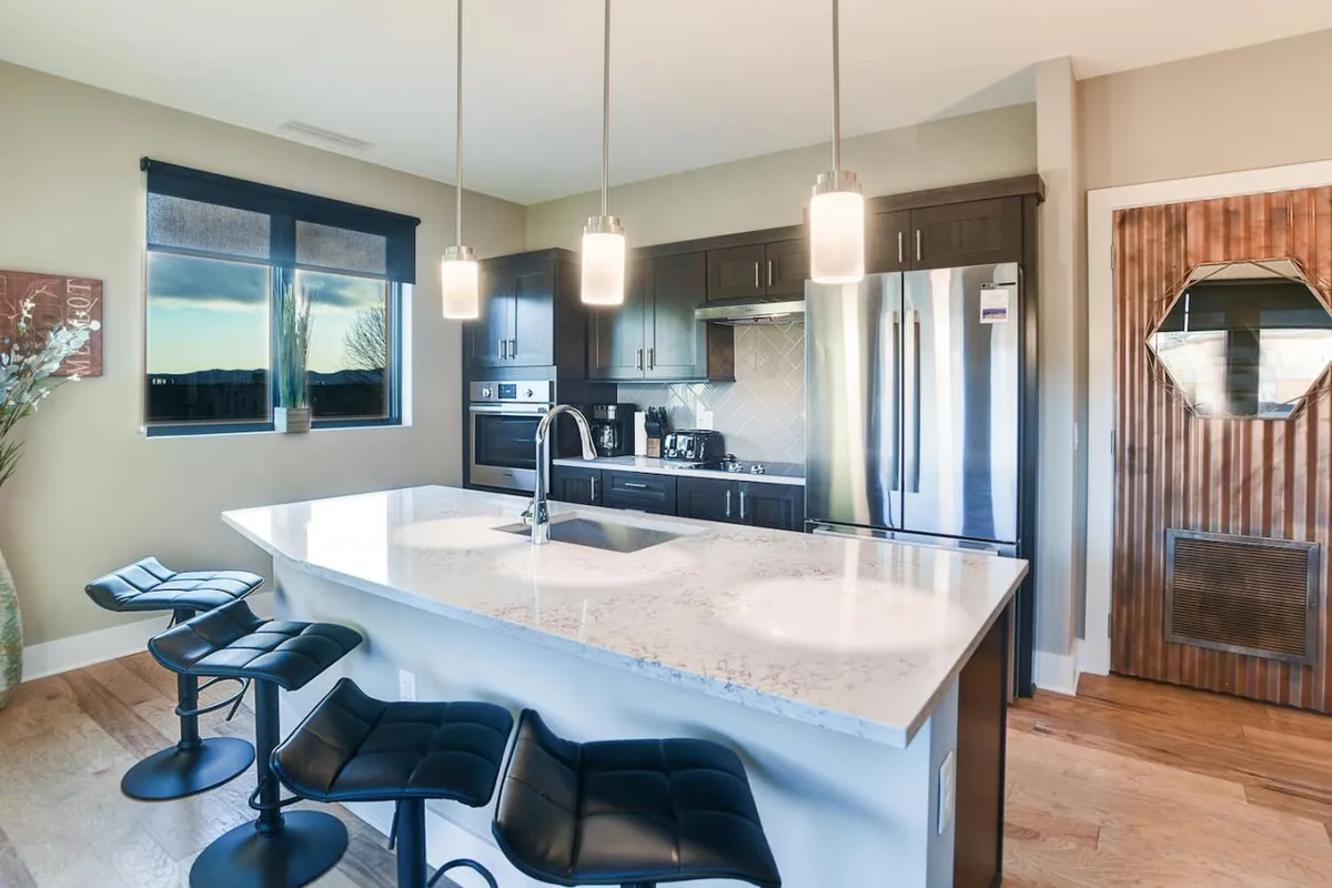 A sunlit kitchen and dining area with a central island, bar stools, modern appliances, and a generous window. The scene radiates a welcoming, contemporary vibe ideal for both quick breakfasts and intimate dinners.